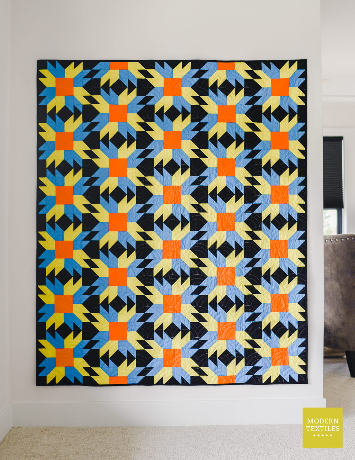 A colorful throw size quilt used as wall hanging with a geometric Sunfire pattern in black, blue, green, and orange, displayed on a white wall in a room with brown chair in background