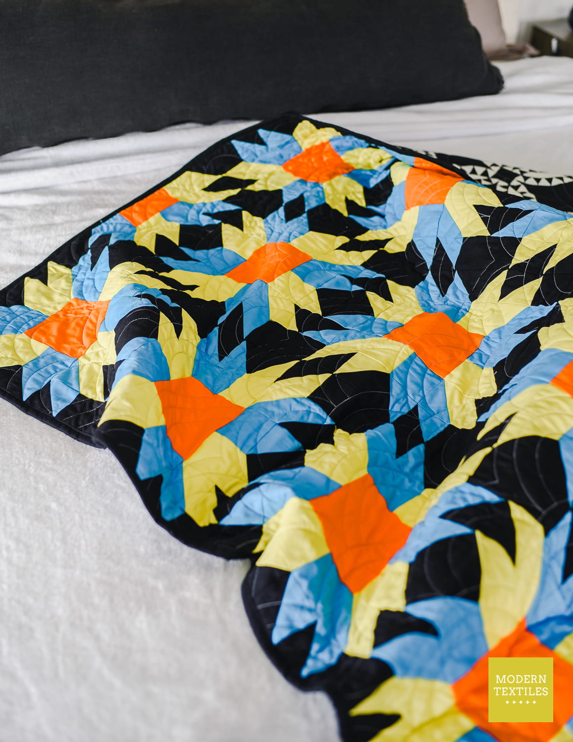 A colorful quilt with a geometric starburst pattern in black, blue, green, and orange, draped over a bed with white sheets and black pillow.