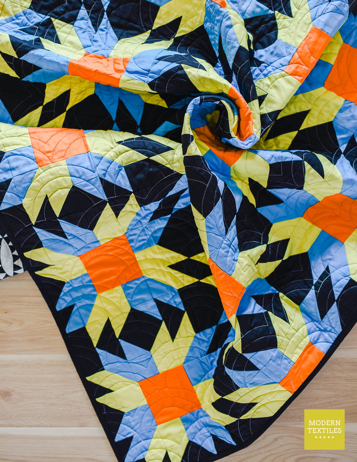 A colorful quilted blanket with a geometric starburst pattern in black, blue, green, and orange, swirled on a light wood floor. 
