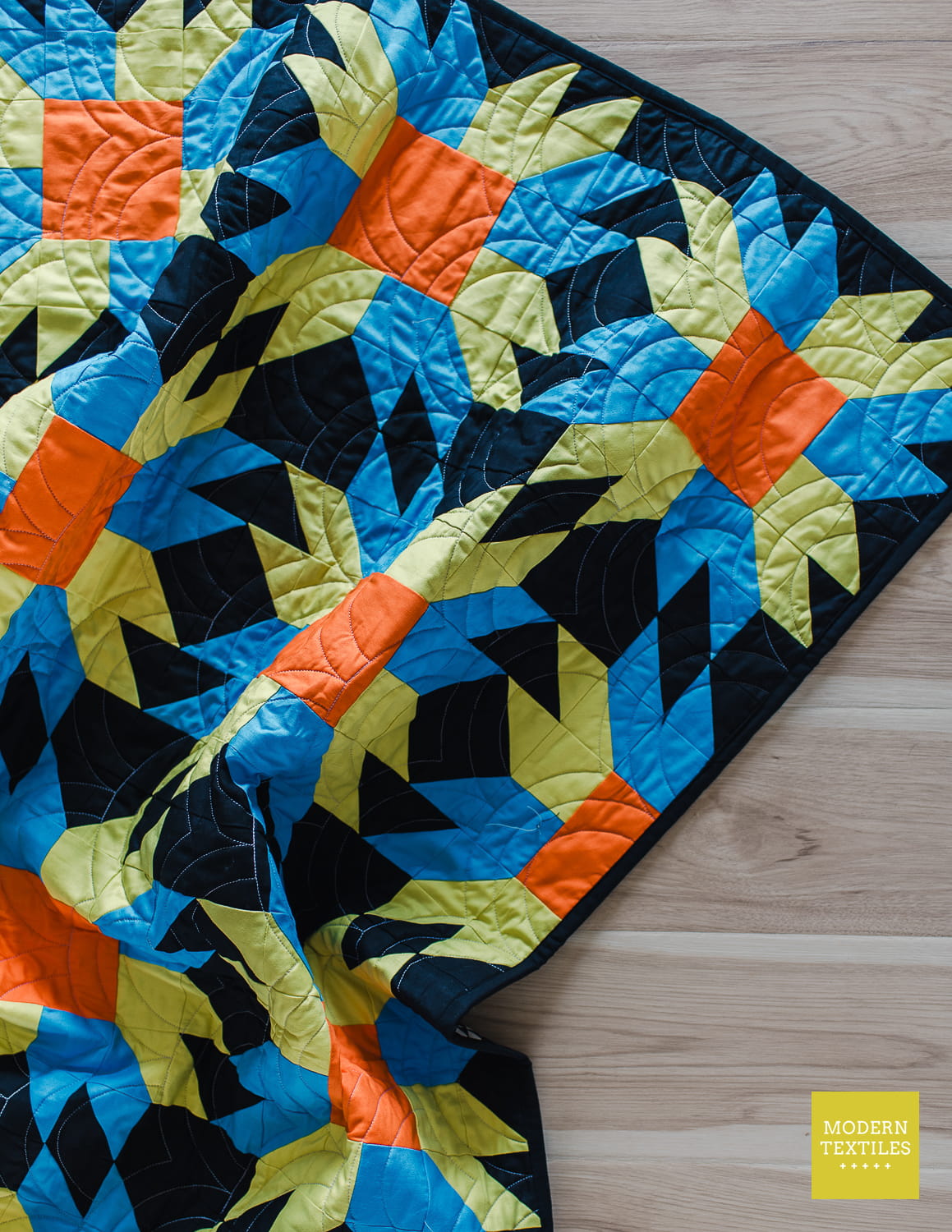 A colorful throw quilt crinkled with a geometric Sunfire pattern in black, blue, green, and orange patchwork, displayed on a natural oak wood floor.
