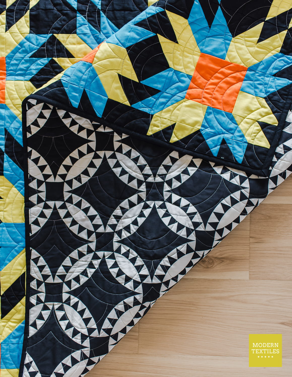 A folded quilted textile blanket or throw is shown on a light wood floor, displaying both its colorful Sinfire pattern in blue, green, and orange, and black patchwork with white and black geometric backing fabric shown.