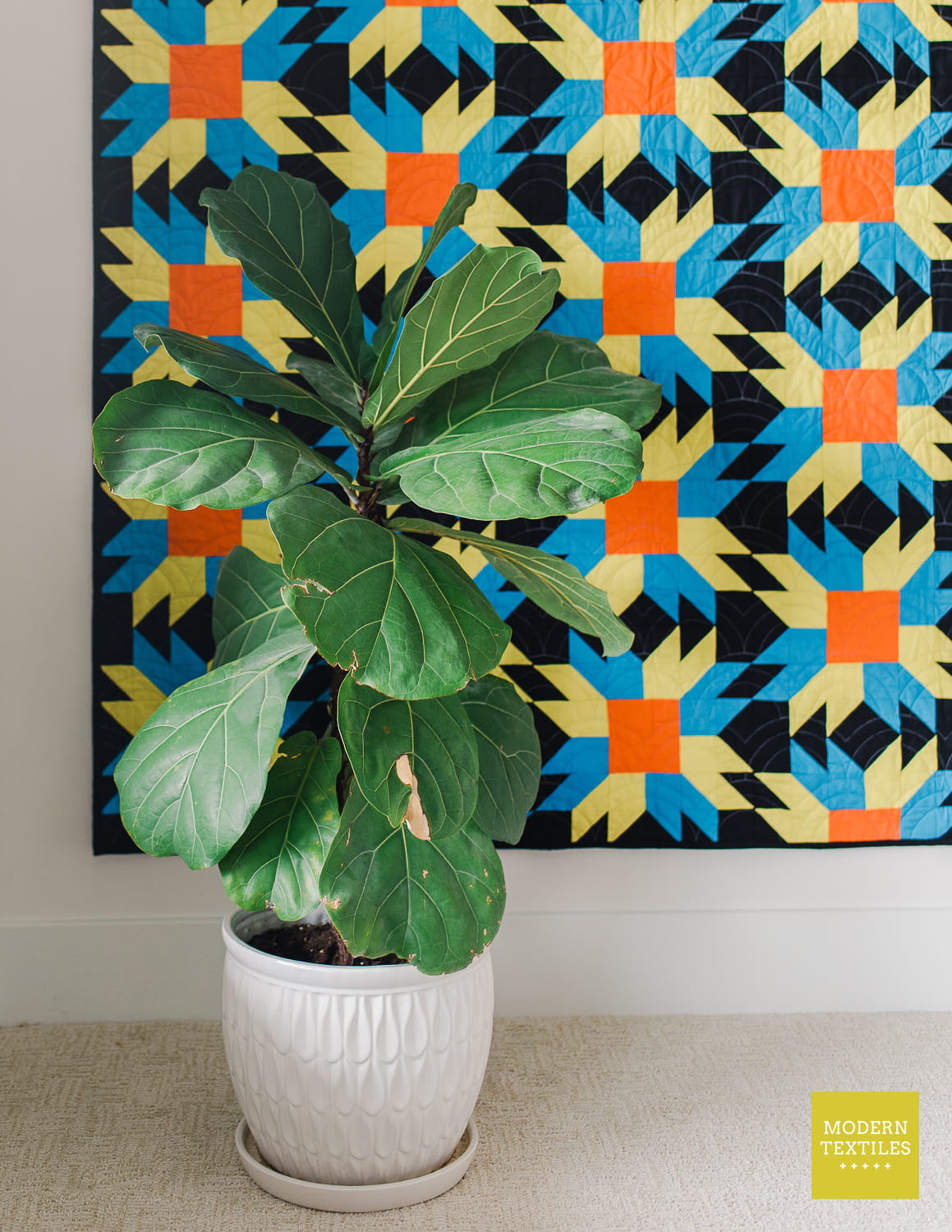 A potted fiddle-leaf fig plant in a white textured ceramic pot, placed in front of a colorful quilt hanging on a wall. The quilt features a geometric sunfire patchwork pattern in black, green, blue, and orange.