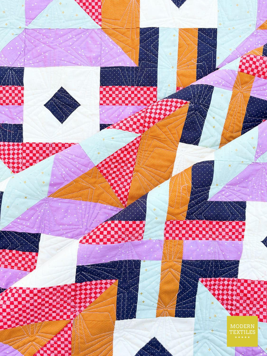 A close-up of a geometric patchwork quilt with blocks of lavender, navy, white, coral red, and orange fabrics, featuring stitched quilting patterns. A small yellow 'Modern Textiles' logo is visible in the bottom right corner.
