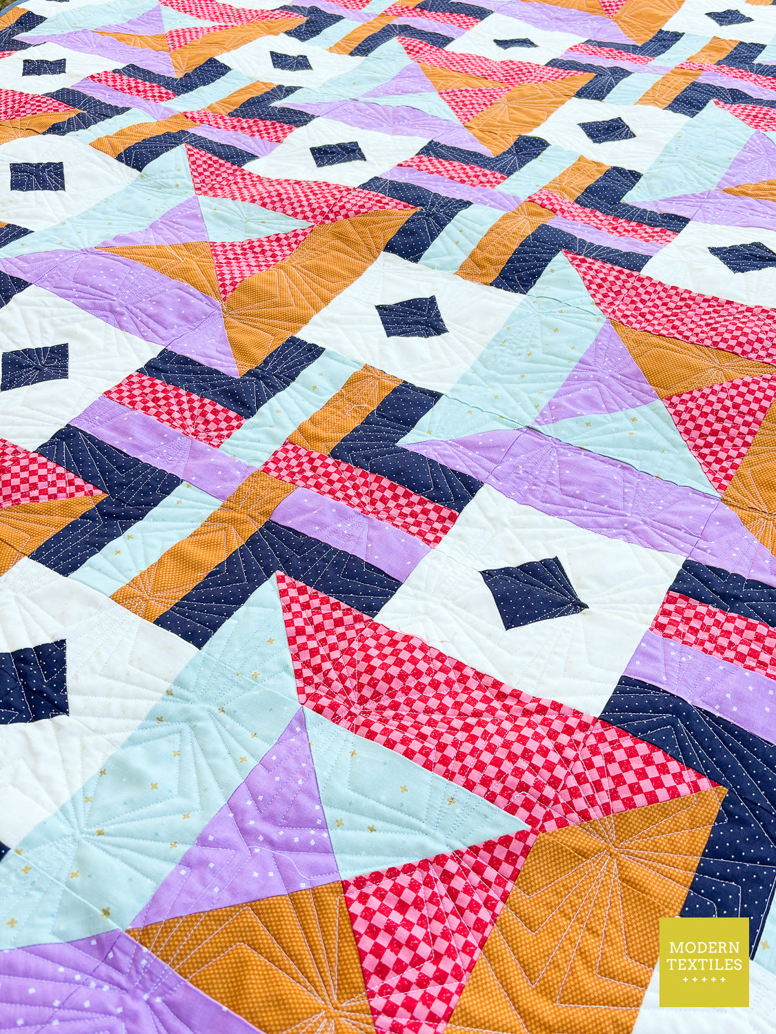 A close-up of a colorful geometric quilted textile with repeating triangles and diamonds in navy, lavender, orange, pink, white, and pale blue, including gingham and polka-dot fabric patterns. A small yellow 'Modern Textiles' label is visible in the lower right corner.