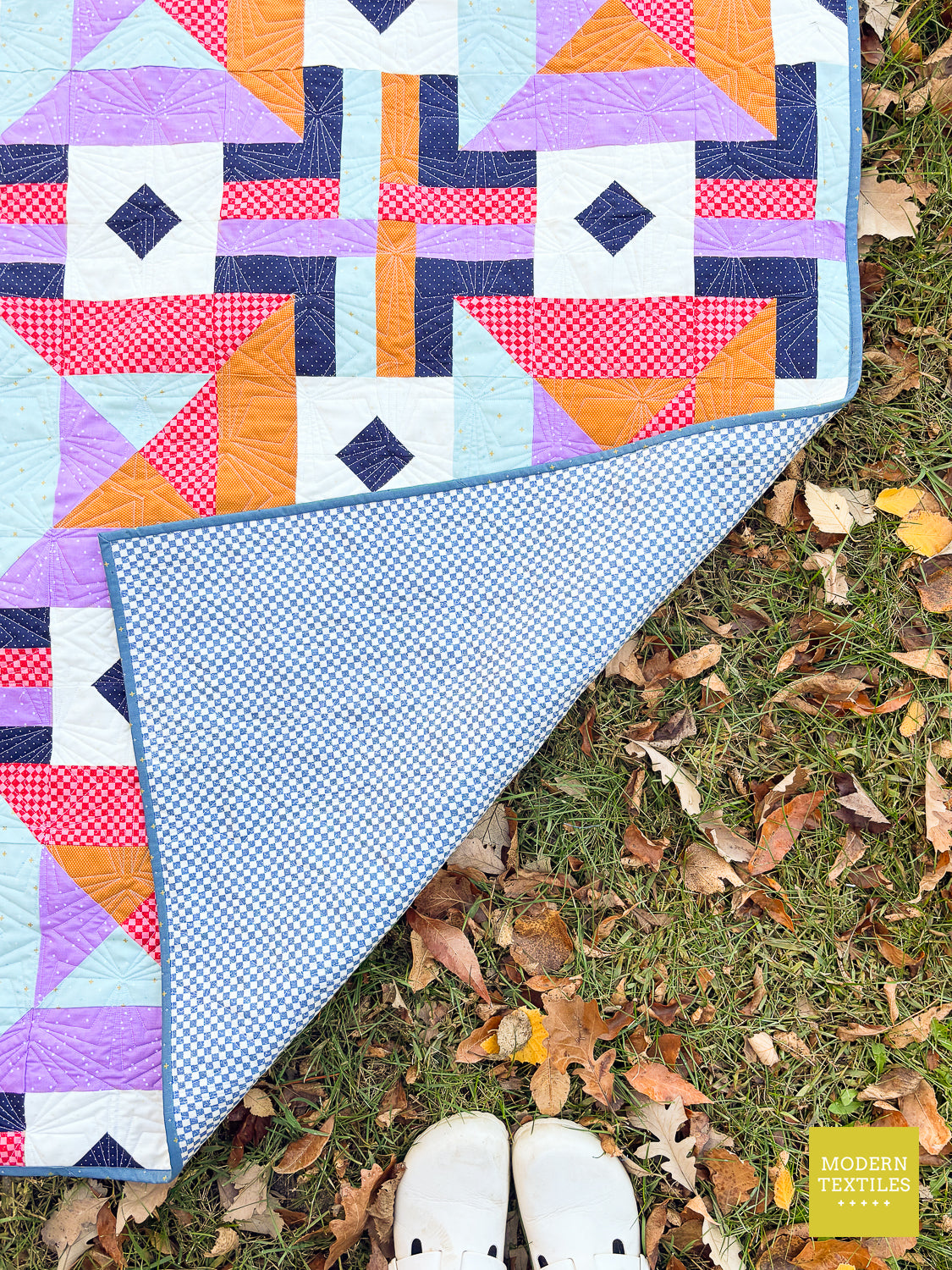 A colorful quilt is draped over grass with scattered autumn leaves, with a person's white shoes visible at the bottom. The quilt features geometric patterns in purple, orange, pink, navy, and white, with a light blue gingham reverse side.