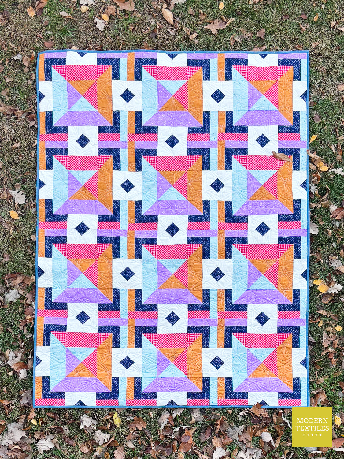 A colorful geometric quilt laid flat on grass with scattered autumn leaves. The quilt features a repeating pattern of pink, orange, purple, light blue, and white blocks framed by navy and purple borders.