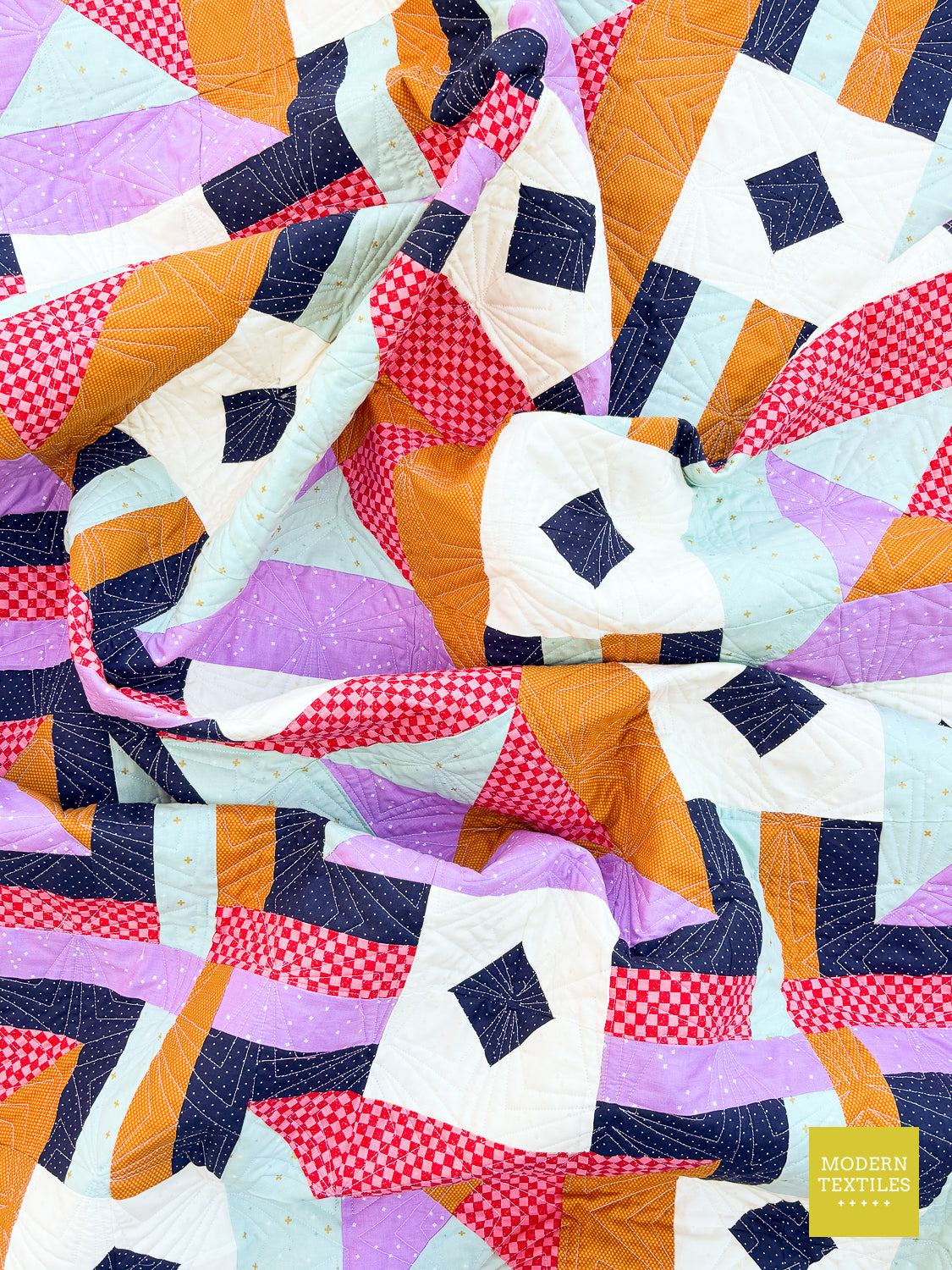 A close-up of a colorful patchwork quilt with geometric blocks in orange, lavender, white, navy, red gingham, and pale blue, with visible quilting lines and a small 'Modern Textiles' logo in the lower right corner.