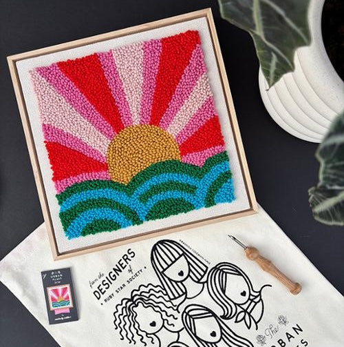 Colorful sun and wave needle punch design in a frame with a plant and stationery items in the background.