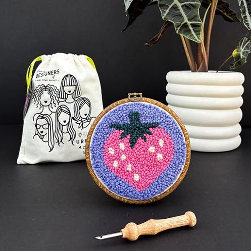Embroidery hoop with pink strawberry and purple background, surrounded by a drawstring bag and crochet hook on a dark surface.